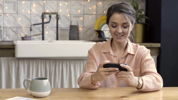 Glimlachend volwassen brunette vrouw met behulp van haar telefoon, spelen spel, zittend in haar keuken aan de houten tafel met een grote mok thee of koffie. Stock footage. Spelen van Smartphone Games, spelletjes verslaving. — Stockvideo