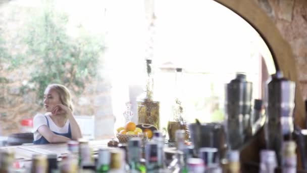 Beautiful blond woman in white t-shirt and denim jumpsuit sitting near bar counter alone and smiling. Art. Young girl with curly hair waiting for someone at bar. — Stock Video