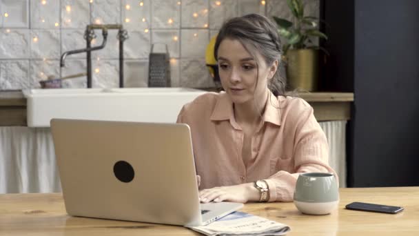 Mujer con una taza grande y su cuaderno en la cocina de su apartamento moderno. Imágenes de archivo. Mujer joven seria sentada en la mesa de la cocina frente a la computadora portátil genérica . — Vídeos de Stock