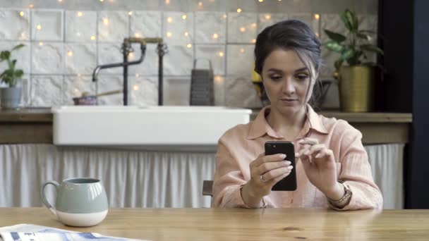 近代的な家でコーヒーマグと台所のテーブルでスマートフォンを使用している若い女性。ストック映像。キッチンのものの背景に彼女のスマートフォンを見て美しいブルネットの女性. — ストック動画