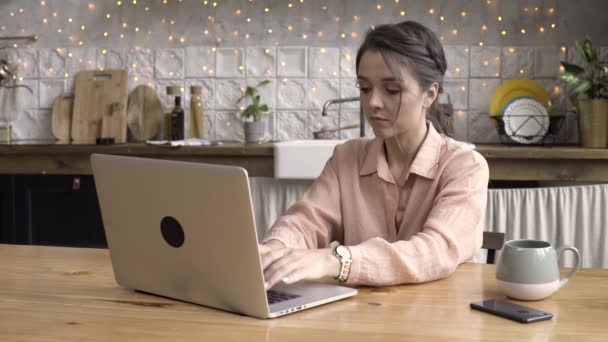 Porträt einer hübschen brünetten Frau, die E-Mails auf dem Laptop schreibt, während sie zu Hause sitzt, freiberufliches Konzept. Archivmaterial. attraktive junge Frau tippt in der Küche auf einem Laptop. — Stockvideo