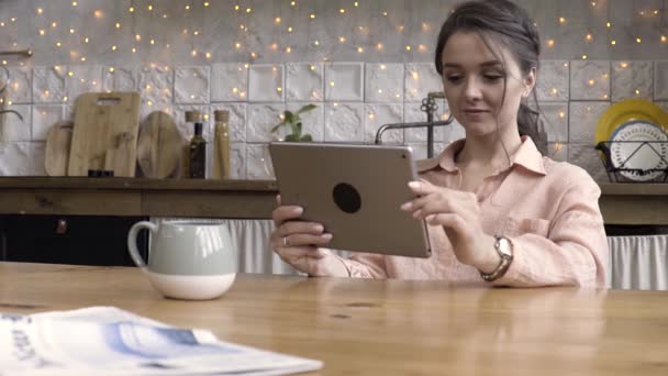 Αξιολάτρευτο και γοητευτικό μελαχρινή γυναίκα κάθεται στο τραπέζι στον τοίχο με τα πράγματα της κουζίνας και λάμπει Γκάρλαντ χρησιμοποιώντας συσκευή tablet. Υλικό από μετοχές. Θηλυκό χρησιμοποιώντας μοντέρνο gadget στο σπίτι. — Αρχείο Βίντεο