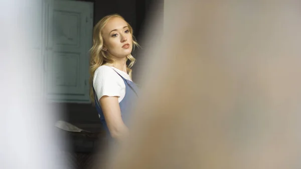 Piękna blondynka w białej koszulce i niebieskim kombinezonie stojąca w salonie, widok zza rozmytej beżowej półki. Sztuka. Przemyślana młoda kobieta w mieszkaniu. — Zdjęcie stockowe