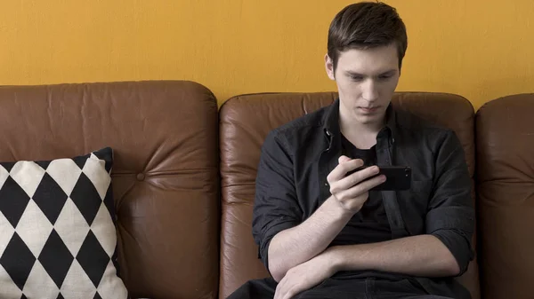Nuori komea mies istuu nahkasohvalla matkapuhelin kotona ja katsoo videota. Varastomateriaalia. Muotokuva miehen ruskea sohva hänen moderni laite . — kuvapankkivalokuva