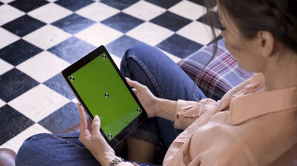 Manos de mujer usando tableta con pantalla verde, sentado en el sofá en casa, vista desde la parte posterior. Imágenes de archivo. Morena joven mujer mirando la tableta con croma key . — Foto de Stock