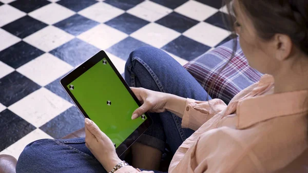 Woman at home relaxing and looking at her tablet computer with green screen on the floor of black and white color. Stock footage. Black tablet with chroma key in the hands of a brunette girl.