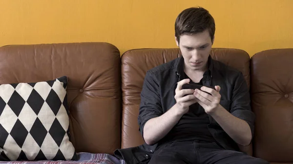 Jeune homme assis sur un canapé en cuir et jouant à des jeux sur son téléphone à la maison sur fond de mur de couleur moutarde. Images d'archives. Homme utilisant son appareil pour jouer à des jeux à la maison . — Photo