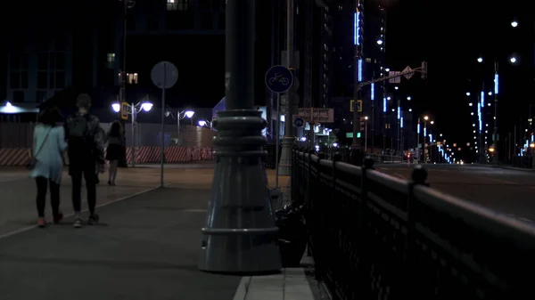 People walking on evening city street along the road lit by street lamps on black sky background. Stock footage. Night view of the city street with vehicles and passers by.