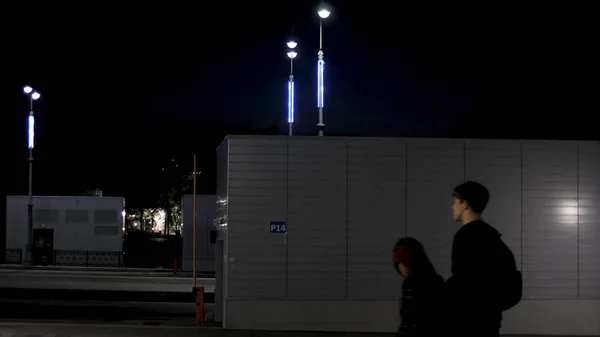 Una pareja caminando por la noche durante el verano en la calle cerca de la carretera vacía iluminada por luces azules de neón. Imágenes de archivo. Adolescentes que pasan por el garaje con barrera . —  Fotos de Stock