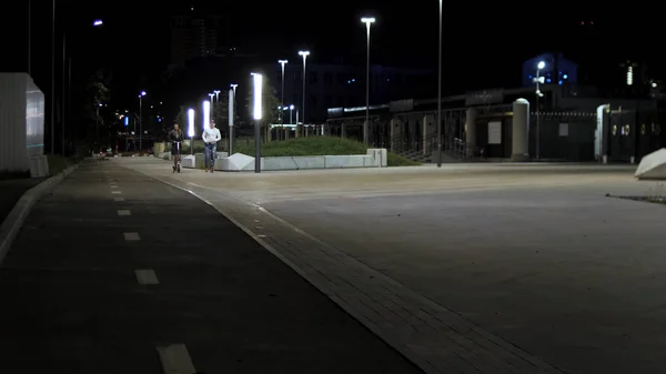 Jovens adolescentes, mulher e homem montando scooters no parque da cidade à noite. Imagens de stock. Casal andando no parque ao longo das luzes da rua e gramado verde . — Fotografia de Stock