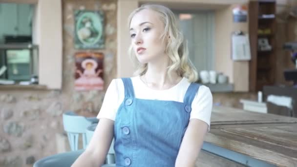Beautiful blond woman in white t-shirt and denim jumpsuit sitting near bar counter alone and looking sad. Art. Young girl with curly hair waiting for someone at bar. — Stock Video