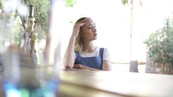 En ung vacker blond kvinna med lockigt hår sitter i baren utanför och tittar åt sidan med huvudet lutade på hennes hand. Artikel. Tankeväckande tjej på bardisken på en sommardag. — Stockvideo