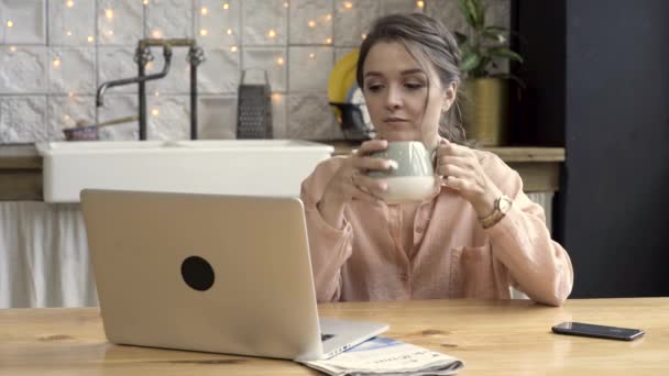 Mujer trabajando desde casa usando laptop en la cocina. Imágenes de archivo. Linda mujer morena bebiendo té de gris y beige taza, trabajando en el ordenador portátil, concepto independiente . — Vídeos de Stock