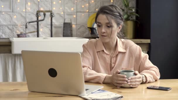 Vuxen mörkhåriga kvinnan värmer händerna med hjälp av varm kaffekopp medan du arbetar på laptop i köket hemma. Stock film. Modern teknik och inredningskoncept. — Stockvideo
