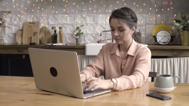 Mujer con una taza grande y su cuaderno en la cocina de su apartamento moderno. Imágenes de archivo. Mujer joven seria sentada en la mesa de la cocina frente a la computadora portátil genérica . — Vídeo de stock