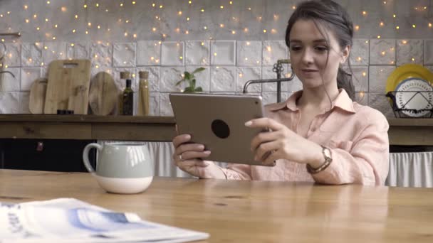 Ritratto di una donna attraente che usa il suo touchpad e legge qualcosa mentre è seduta in cucina contro un muro decorato con cose diverse. Filmati delle scorte. Concetto di tecnologie moderne . — Video Stock