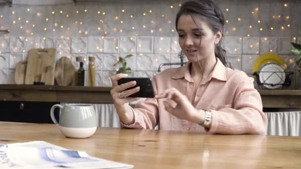 Jovem mulherjogando jogos de smartphone na mesa da cozinha com caneca de café em uma casa moderna. Imagens de stock. Linda morena feminina olhando para seu smartphone no fundo de material de cozinha . — Vídeo de Stock