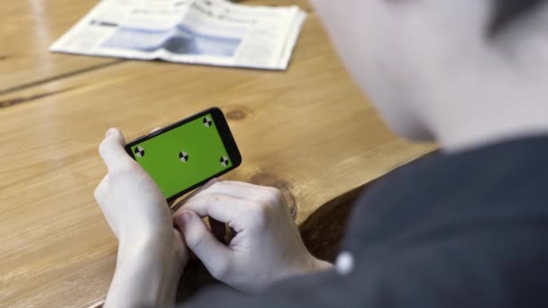 Smartphone orientação horizontal com uma tela verde: tiro sobre o ombro de um jovem. Filmagem de stock. Homem sentado à mesa, tocando e rolando no cromo do telefone . — Vídeo de Stock