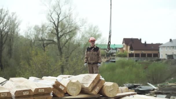 Budowniczy noszący mundur i kask ochronny stojący i patrzący na ruchomy hak dźwigu na placu budowy. Klip. Hak przemysłowy i proca do ciężkich obciążeń i wielu drewnianych belek. — Wideo stockowe