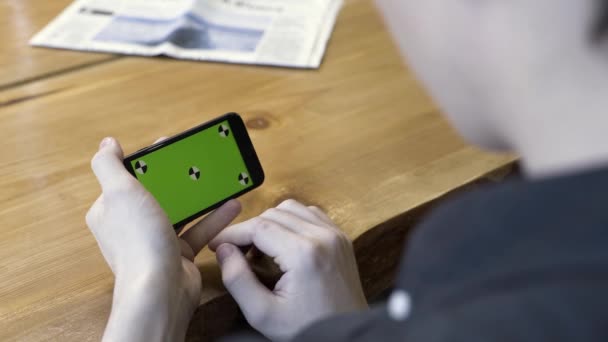 Orientación horizontal del smartphone con pantalla verde: disparo sobre el hombro de un joven. Imágenes de archivo. Hombre sentado en la mesa, tocando, desplazándose y acercándose al cromakey del teléfono . — Vídeos de Stock