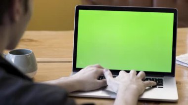 Laptopta yeşil ekranla yazı yazan bir adamın arka görüntüsü, büyük bir fincan çay veya kahveyle ahşap masada oturuyor. Stok görüntüleri. Bilgisayarda masasında çalışan bir erkek, kromakey..