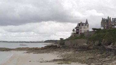 Sahilde, tuğla kulübeleri ve arka planda mavi bulutlu gökyüzü olan taşlı sahilde yürüyen bir grup insan için hava. Başla. Kumlu sahilde yürüyen arkadaşların yan görüntüsü.