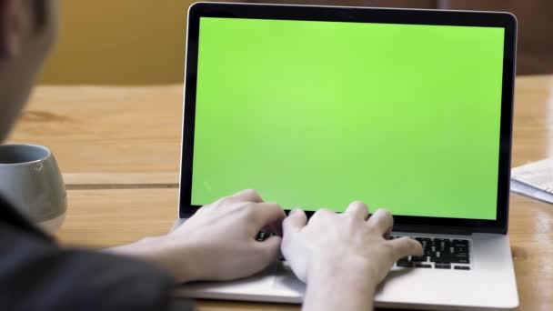 Achteraanzicht van een man die typt op de laptop met groen scherm, zittend aan de houten tafel met een grote kop thee of koffie. Voorraadbeelden. Man werkt aan zijn bureau aan de computer, chromakey. — Stockvideo