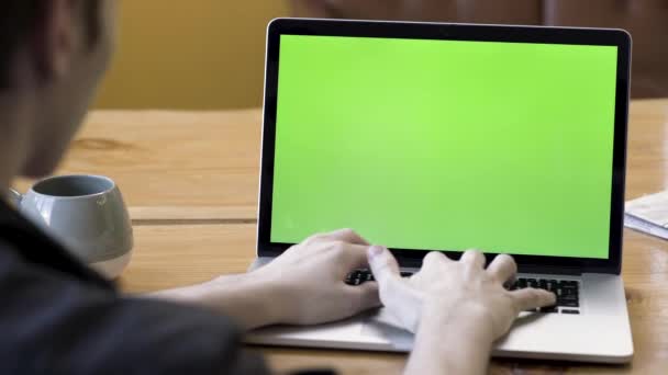 お茶やコーヒーの大きなカップで木製のテーブルに座って、緑の画面でノートパソコンに入力する男のリアビュー。ストック映像だ男はコンピューターの机で働いてるクロマキー. — ストック動画