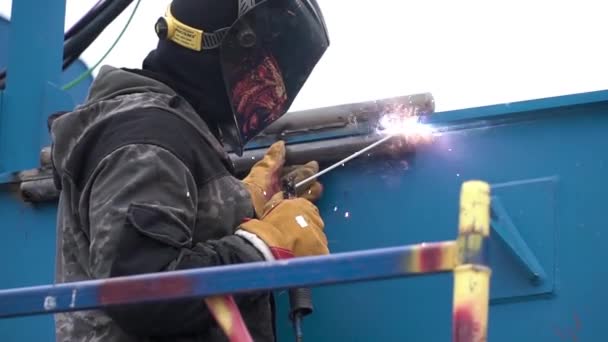 Vista lateral de um homem em máscara de solda protetora enquanto solda objetos de metal azul processo no local de construção. Clipe. Trabalhador usando calor alto para derreter as peças de metal juntos . — Vídeo de Stock