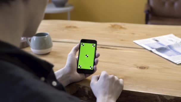 녹색 스크린 크롬 키를 장착 한 무기 전자 기기를 들고 있는 한 남자가 나무 탁자 위에서 스크롤하고 두드리고 있습니다. 주가 영상이요. 사람이 앉아서 휴대 전화를 사용하는 방. — 비디오