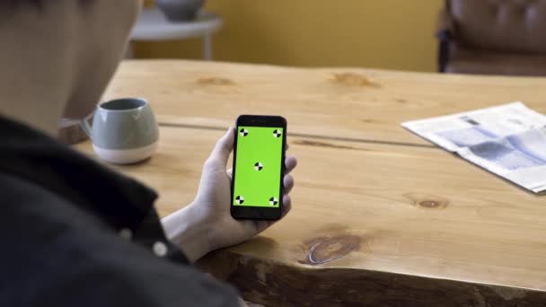 Προβολή πάνω από τον ώμο ενός επιχειρηματία χρησιμοποιώντας το κινητό τηλέφωνο χειρός με πράσινη οθόνη touchpad σε ξύλινο τραπέζι φόντο. Στικ. Ο άνθρωπος κοιτάζει τη συσκευή του, chromakey. — Αρχείο Βίντεο