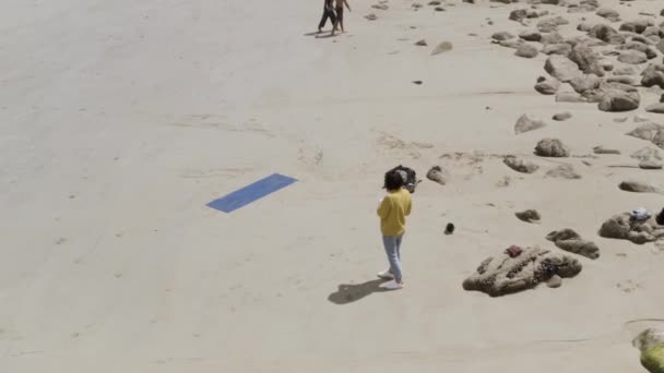 Flygfoto av en kvinna i gul tröja och blå jeans som står på sandstranden nära sin yogamatta. Börja. Kvinna stående på sanden nära stenarna. — Stockvideo