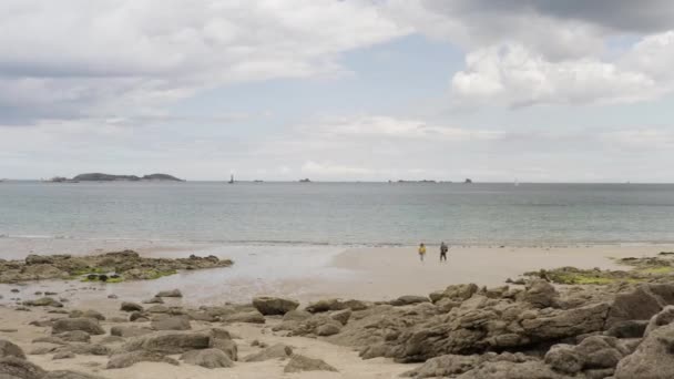 Par går på stranden med stenig strand med lugnt hav och blå molnig himmel på bakgrunden. Börja. Man och kvinna går på sandkusten. — Stockvideo