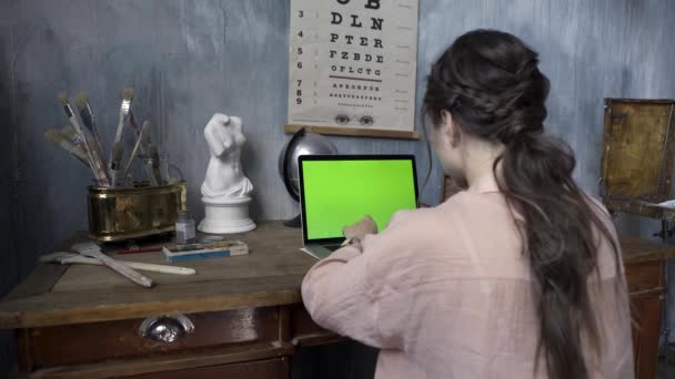 Sobre el hombro de una mujer escribiendo en una computadora portátil con una pantalla verde cromakey. Imágenes de archivo. Manos de mujer escribiendo en el portátil con pantalla verde en el taller de artista . — Vídeo de stock