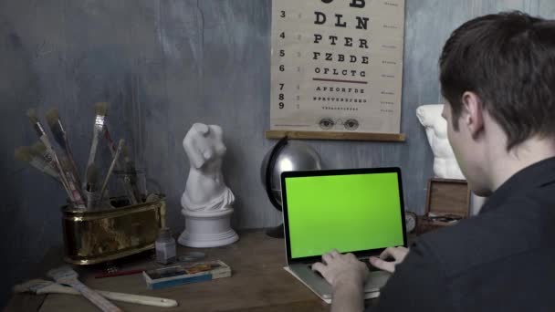 Visão traseira do artesão masculino trabalhando na mesa no laptop com chromakey na oficina do artista. Imagens de stock. Homem digitando no computador com tela verde na sala decorada . — Vídeo de Stock