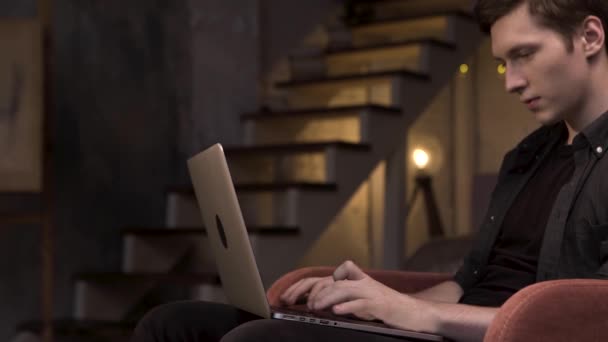 Zijaanzicht van de jongeman die laptop gebruikt terwijl hij thuis op de achtergrond van de trap zit. Voorraadbeelden. Man zit in de rode fauteuil en typt op zijn computer in de schemerkamer. — Stockvideo