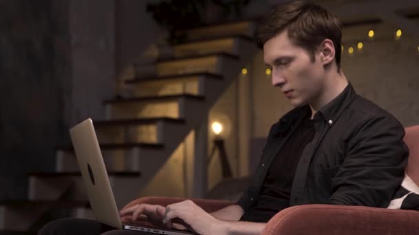 Πλευρική άποψη του νεαρού άνδρα που χρησιμοποιεί φορητό υπολογιστή, ενώ κάθεται στο σπίτι στις σκάλες φόντο. Στικ. Ο άνθρωπος κάθεται στην κόκκινη πολυθρόνα και πληκτρολογεί στον υπολογιστή του στο σκοτεινό δωμάτιο. — Αρχείο Βίντεο