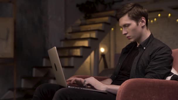 Komoly jóképű, fekete inges szabadúszó, aki laptoppal dolgozik, miközben a piros karosszékben ül, kezével a billentyűzeten. Készletfelvétel. Fiatal férfi használ számítógépet a lépcsőn a háttérben. — Stock videók