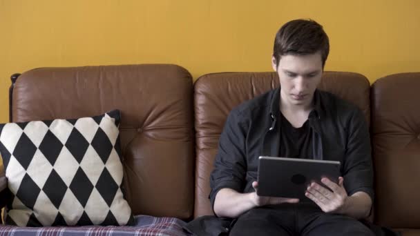一个笑着用平板电脑的年轻人坐在公寓里的棕色皮沙发上的画像。 库存录像。 男人在芥末色墙的背景上敲敲他的装置屏幕. — 图库视频影像