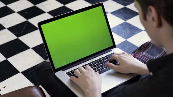 초록색 화면을 가진 컴퓨터를 사용하고 편안 한 소파에 앉아 있는 한 남자의 어깨 너머의 모습. 주가 영상이요. 백층 배경에 크로마키를 가지고 노트북으로 타자 하는 사업가. — 비디오