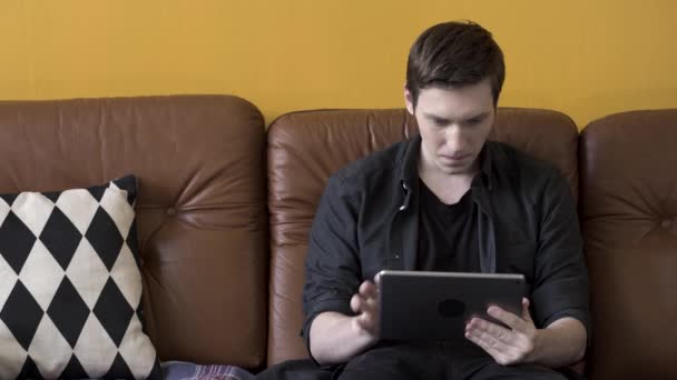 坐在家里的棕色皮沙发上，穿着黑色衬衫的紧张男人在用他的平板电脑。 库存录像。 穿着黑色衣服，手持现代触摸板装置的时髦博客. — 图库视频影像