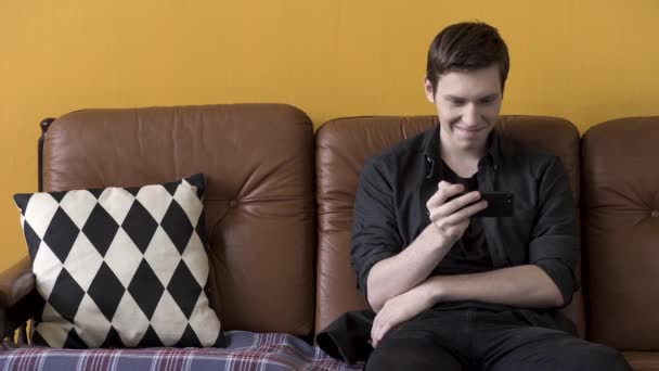 Τεχνολογία έννοια, ο άνθρωπος βλέποντας αστείο βίντεο στο smartphone και κάθεται σε ένα άνετο καφέ δερμάτινο καναπέ με μαύρο και άσπρο μαξιλάρι. Στικ. Νεαρός που χρησιμοποιεί σύγχρονη συσκευή. — Αρχείο Βίντεο