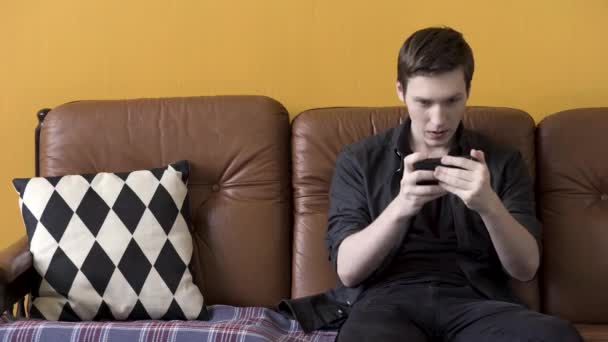 Concept technologique, homme jouant au jeu sur smartphone et assis sur un canapé en cuir marron confortable avec oreiller noir et blanc. Images d'archives. Jeune homme utilisant un appareil moderne . — Video