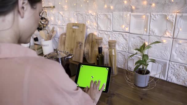 Vista trasera de una mujer tocando en una tableta con pantalla verde en la cocina en casa para recetas. Imágenes de archivo. Mujer usando su tableta con croma key para cocinar . — Vídeos de Stock