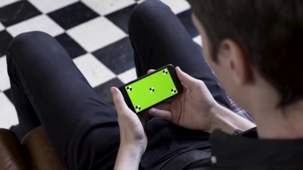 チェックされた黒と白の床の背景にソファに座っている間、緑の画面を持つスマートフォンを使用してビジネスマン。ストック映像だ水平位置にデバイスを保持し、見て男. — ストック動画