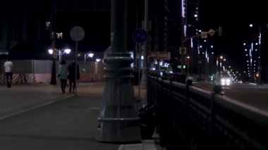 Gece şehir caddesinde yürüyen insanlar kara gökyüzü arka planında sokak lambalarıyla aydınlatılıyor. Stok görüntüleri. Şehir caddesinin gece görüş alanında araçlar ve yoldan geçenler var..