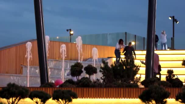 Les gens assis et debout sur les grands escaliers lumineux dans le parc de la ville en face de la fontaine. Images d'archives. Hommes et femmes passent du temps dans les lieux publics avec une architecture moderne . — Video