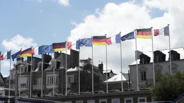 Ευρωπαϊκές σημαίες ταλαντεύεται στον άνεμο με όμορφα σπίτια και μπλε, συννεφιασμένο ουρανό στο παρασκήνιο. Στικ. Σημαίες της Γερμανίας, της Γαλλίας και της Ευρωπαϊκής Ένωσης μπροστά από τα σπίτια. — Αρχείο Βίντεο
