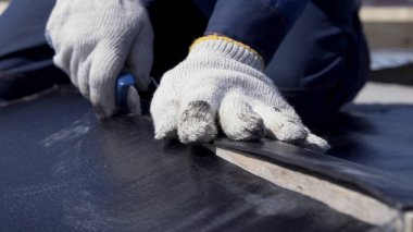 Siyah ruberoid tabakasını teknik kesici ile kesen beyaz koruyucu eldivenli işçi elleri için yakın çekim. Şarjör. İşçi, çatı malzemelerinden bir parça kesiyor..