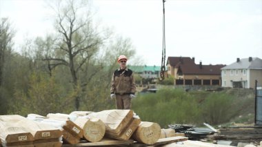 Üniformalı ve koruyucu başlıklı adam inşaat alanındaki vinç kancasına bakıyor. Şarjör. Ağır yük için sanayi kancası ve askı ve bir sürü tahta kiriş..